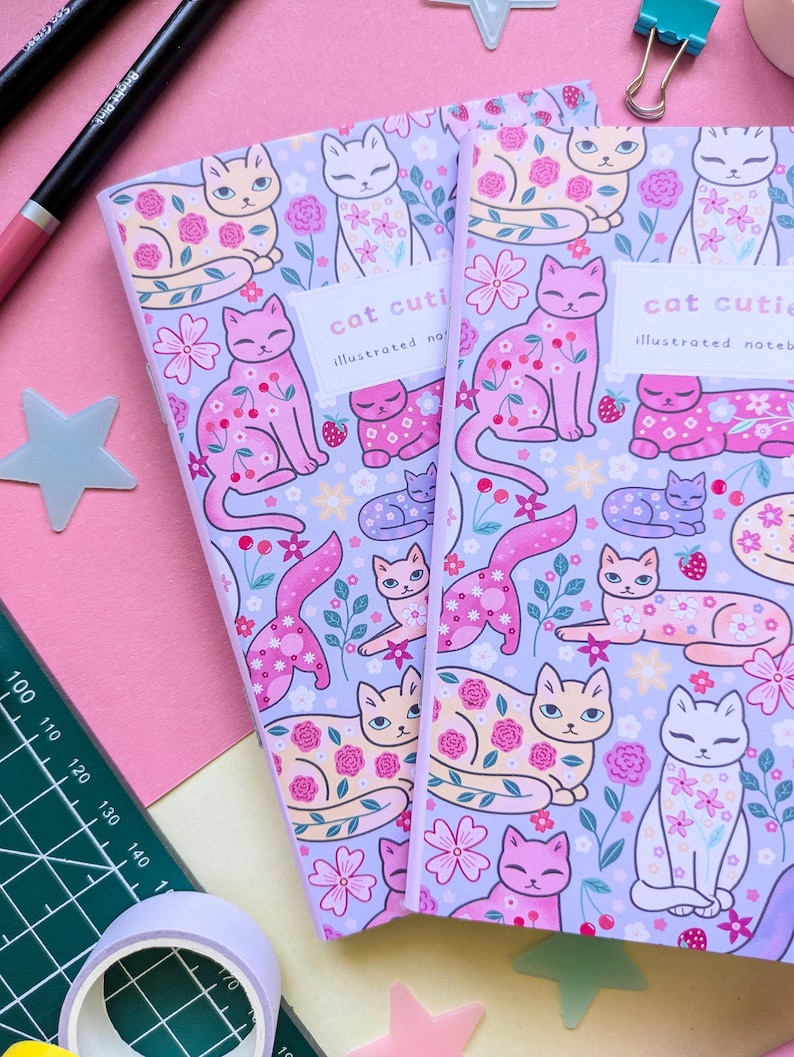 May include: Two notebooks with a lavender background and a repeating pattern of cartoon cats, flowers, and strawberries. The notebooks have the text "cat cutie" and "illustrated notebook" on the cover. Other stationery items, including pens, tape, and paperclips, are scattered around the notebooks.