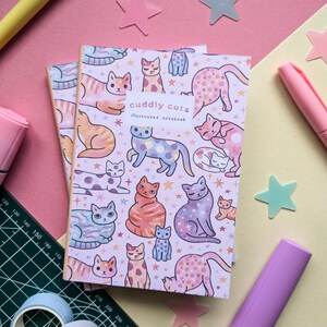 May include: A pink and white illustrated notebook with a pattern of colorful cats and stars. The notebook cover says "cuddly cats illustrated notebook".