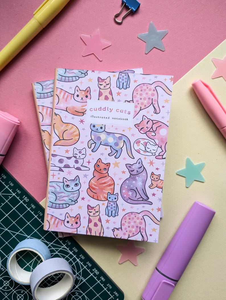 May include: A pink and white illustrated notebook with a pattern of colorful cats and stars. The notebook cover says "cuddly cats illustrated notebook".