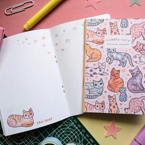 May include: A pink and white illustrated notebook featuring a cat design. The cover showcases a range of colourful cats with various patterns and poses. The text on the cover reads "Cuddly Cats Illustrated Notebook".