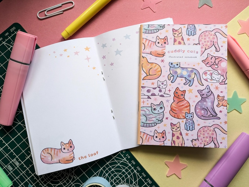 May include: A pink and white illustrated notebook with a cat design. The cover features a variety of colorful cats with different patterns and poses. The text on the cover reads "Cuddly Cats Illustrated Notebook".