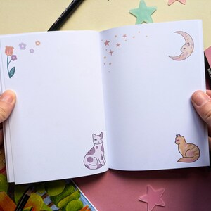 May include: An open notebook with a white background and colorful illustrations of flowers, stars, a crescent moon, and cats. The notebook is perfect for journaling, sketching, or taking notes.