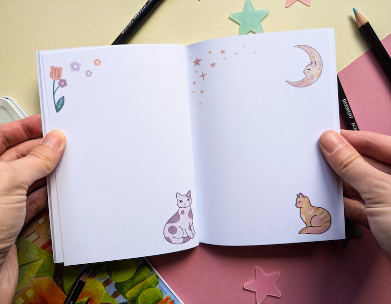 May include: An open notebook with a white background and colorful illustrations of flowers, stars, a crescent moon, and cats. The notebook is perfect for journaling, sketching, or taking notes.