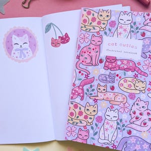 May include: An open notebook with blank pages next to a closed notebook. The closed notebook has a lavender cover with illustrations of cats, flowers, and strawberries. The text "cat cuties illustrated notebook" is on the cover. A pink background.