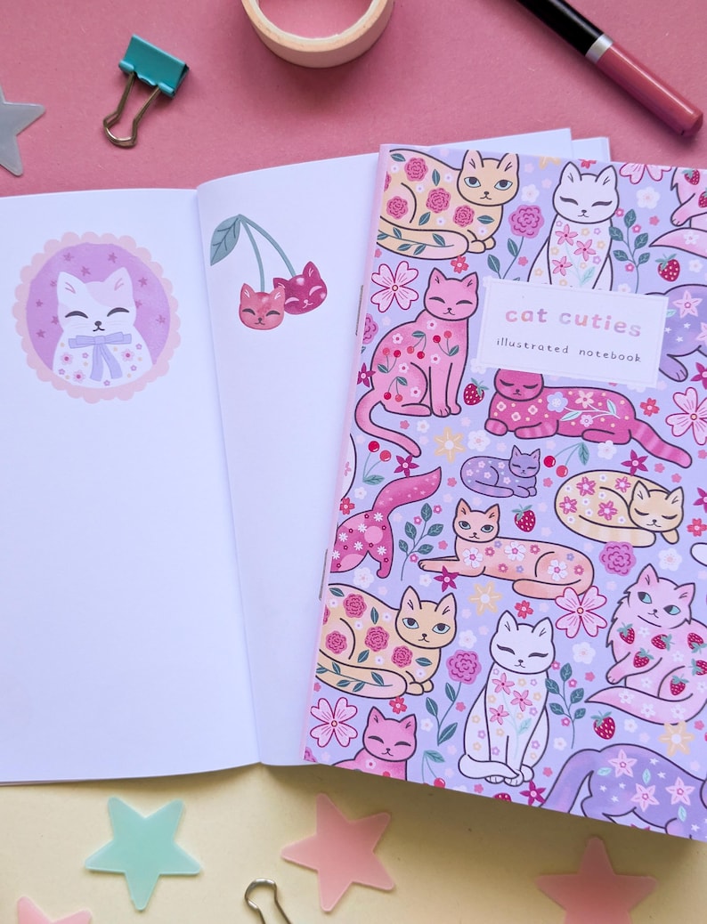 May include: An open notebook with blank pages next to a closed notebook. The closed notebook has a lavender cover with illustrations of cats, flowers, and strawberries. The text "cat cuties illustrated notebook" is on the cover. A pink background.