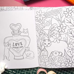 K&ouml;nnte beinhalten: Offene Seiten eines Malbuchs mit Katzenillustrationen. Eine Seite zeigt einen Flakon mit der Aufschrift "LOVE PURRTION" und Katzen. Die andere Seite zeigt eine Katze in einem Blumengarten. Bleistifte und eine Schneidematte sind sichtbar.