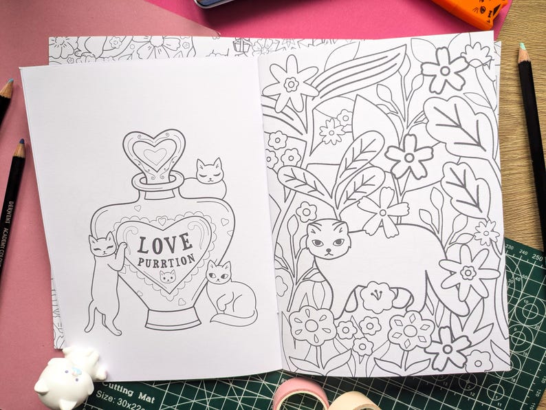 K&ouml;nnte beinhalten: Offene Seiten eines Malbuchs mit Katzenillustrationen. Eine Seite zeigt einen Flakon mit der Aufschrift "LOVE PURRTION" und Katzen. Die andere Seite zeigt eine Katze in einem Blumengarten. Bleistifte und eine Schneidematte sind sichtbar.