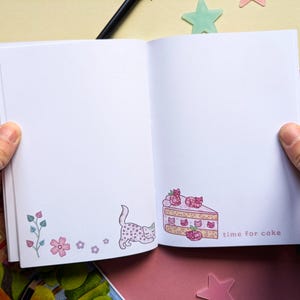 May include: Open notebook with a pink and white cake slice illustration on the right page and a pink and white cat illustration on the left page. The text "time for cake" is written below the cake slice.