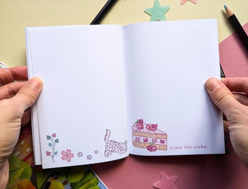May include: Open notebook with a pink and white cake slice illustration on the right page and a pink and white cat illustration on the left page. The text "time for cake" is written below the cake slice.