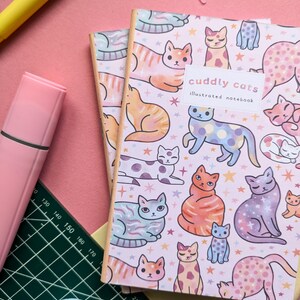May include: Two pink and white illustrated notebooks with a pattern of colorful cats and stars. The front cover of the top notebook says "cuddly cats illustrated notebook".