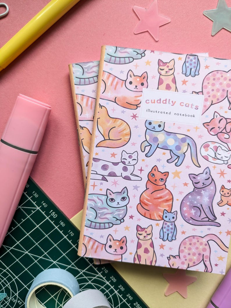 May include: Two pink and white illustrated notebooks with a pattern of colorful cats and stars. The front cover of the top notebook says "cuddly cats illustrated notebook".