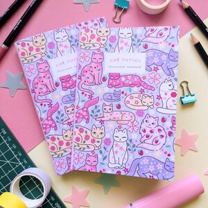 May include: Two notebooks with a lavender cover featuring a pattern of illustrated cats, flowers, and berries. The notebooks have the text "cat cuties illustrated notebook". The background includes pink and yellow stationery items.