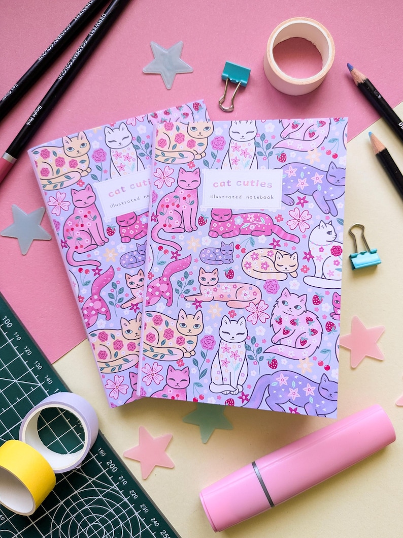 May include: Two notebooks with a lavender cover featuring a pattern of illustrated cats, flowers, and berries. The notebooks have the text "cat cuties illustrated notebook". The background includes pink and yellow stationery items.