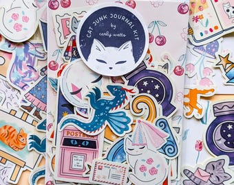 May include: A Cat Junk Journal Kit by Carly Watts in a clear plastic sleeve. The kit contains various die-cut stickers featuring cats, a blue dragon, a pink mailbox, a crescent moon, and cherry patterns. A circular label with a white cat illustration and the text Cat Junk Journal Kit by Carly Watts is centered on the front.
