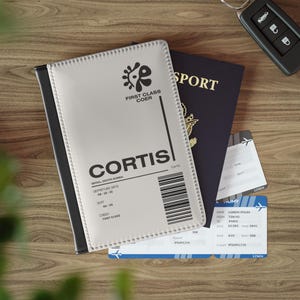 Könnte beinhalten: Ein hellgrauer Reisepasshalter mit dem Text "CORTIS" und "FIRST CLASS COER" liegt neben einem dunkelblauen Reisepass und zwei Flugtickets. Ein Autoschlüssel befindet sich im Hintergrund.