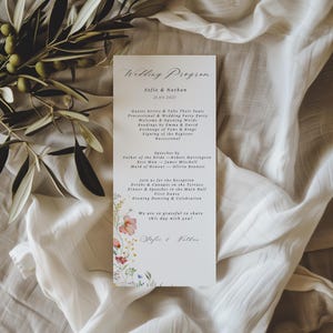 May include: A vertical white wedding programme card featuring black text detailing the ceremony schedule for Sofie and Nathan on 21.03.2027. The bottom left corner is decorated with a colourful watercolour illustration of wildflowers and greenery.