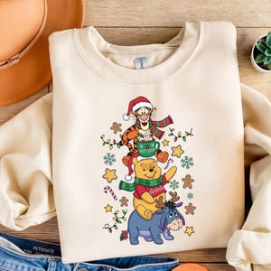 May include: Cream-colored sweatshirt featuring a festive design with Winnie the Pooh characters. Tigger wears a Santa hat and scarf, holding a "Hot Cocoa" mug. Pooh and Eeyore are also dressed for the holidays, surrounded by Christmas lights and gingerbread men.