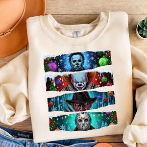 Könnte beinhalten: Cremefarbenes Sweatshirt mit vier Horrorfilmfiguren: Michael Myers, Pennywise, Freddy Krueger und Jason Voorhees. Jede Figur ist vor einem winterlichen Hintergrund mit Weihnachtsschmuck und Schneeflocken platziert.