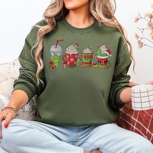 May include: An olive green sweatshirt featuring a festive design of four holiday-themed beverages. Each drink has a Grinch character and holiday decorations. The sweatshirt is a long-sleeved crew neck.