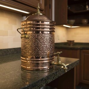 May include: A copper water dispenser with an ornate, embossed design. The dispenser has a lid, handles, and a brass spigot. It is displayed on a dark countertop in a kitchen setting.