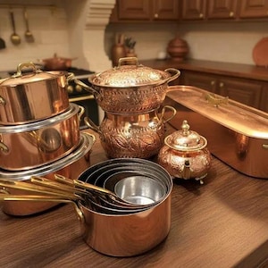 Puede incluir: Colección de utensilios de cocina de cobre, que incluye ollas, sartenes y tazas medidoras, dispuestas sobre una superficie de madera. Las ollas tienen asas y tapas de latón. El cobre tiene una superficie brillante.