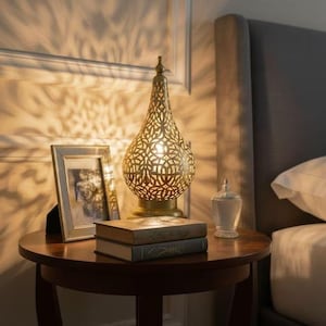 Puede incluir: Una lámpara de mesa dorada y ornamentada con un diseño calado se encuentra sobre una mesa de madera oscura. Una foto enmarcada y dos libros también están sobre la mesa. La lámpara proyecta una luz estampada en la pared. Un tarro de cristal con tapa también está sobre la mesa.