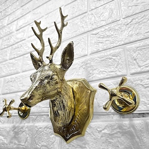 Puede incluir: Un soporte de pared con cabeza de ciervo de latón con un juego de grifos decorativos. La cabeza de ciervo está montada en una placa en forma de escudo. El juego de grifos incluye dos manijas en forma de cruz. Los accesorios son dorados.