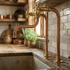 Può includere: Un rubinetto da cucina in rame e ottone dal design vintage. Il rubinetto presenta tubi in rame curvi e maniglie e dettagli in ottone. È montato su un piano di lavoro in legno invecchiato, con un lavandino in pietra in primo piano. Lo sfondo include mensole e una finestra.