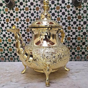 Könnte beinhalten: Eine goldfarbene Teekanne mit aufwendigen geprägten Designs, einem langen Ausguss und einem dekorativen Deckel. Die Teekanne hat drei kleine Füße und einen Griff. Der Hintergrund zeigt eine Mosaikfliesenwand.