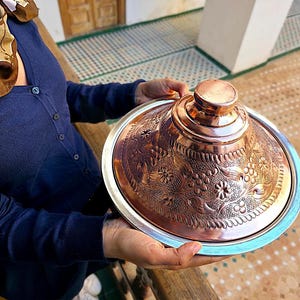 Pode incluir: Um tagine de cobre com intrincados padrões florais e geométricos, segurado por uma pessoa. O tagine apresenta uma tampa cónica e uma base larga com borda prateada. O cobre tem um tom castanho-avermelhado quente.