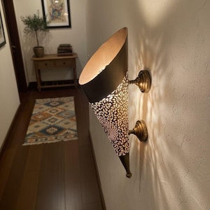 Puede incluir: Aplique de pared decorativo con una pantalla en forma de cono. La pantalla es negra por fuera y dorada por dentro, con un patrón calado detallado. El aplique está montado en la pared con dos fijaciones de color latón.