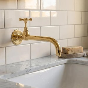 Puede incluir: Un grifo dorado con un mango en forma de cruz está montado en una pared de azulejos blancos. Una pastilla de jabón descansa sobre un pequeño soporte de piedra redondo. Una toalla de lino doblada está sobre la encimera.