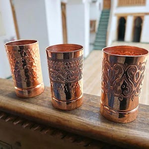 May include: Three copper drinking glasses with detailed patterns. The glasses are cylindrical and made of polished copper. Textures include hammered and engraved designs. The glasses are displayed on a wooden surface.
