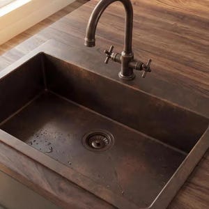 May include: A copper-toned kitchen sink with a matching faucet. The rectangular sink is set into a dark wood countertop. The faucet has cross-shaped handles. The sink is in a kitchen setting.