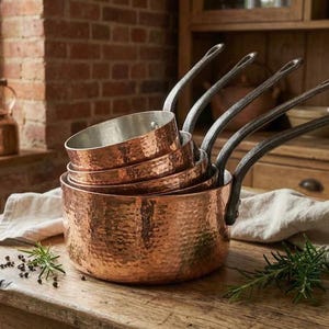 Puede incluir: Un juego de cinco cacerolas de cobre con exteriores martillados y largos mangos de metal oscuro. Las cacerolas están apiladas sobre una superficie de madera con hierbas y granos de pimienta. Una tetera de cobre es visible en el fondo.