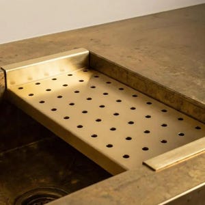 May include: A close-up of a brass sink with a built-in perforated tray. The sink and countertop have a warm, metallic finish. The tray is rectangular with evenly spaced holes.