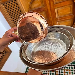 May include: A copper saucepan with a gold handle is held above a stack of hammered metal serving trays. The trays are in varying sizes and have a silver finish. The copper pan and trays have a textured, dimpled surface. The background includes wooden architectural details.