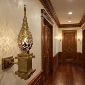 Puede incluir: Aplique de pared de latón con una pantalla en forma de lágrima con un delicado diseño calado. El aplique está montado sobre una base y un brazo cuadrados, proyectando intrincados patrones de luz en la pared. El pasillo tiene paneles de madera y puertas.