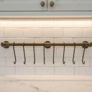 Puede incluir: Un estante de utensilios de cocina de color bronce con siete ganchos, montado en un salpicadero de azulejos blancos. El estante está situado debajo de armarios blancos con herrajes de latón, y una tira de luz ilumina la zona.