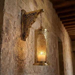 Puede incluir: Una linterna dorada y ornamentada, montada en la pared, con una vela dentro. La linterna está suspendida de un soporte decorativo y una cadena. El intrincado diseño proyecta una sombra estampada sobre la pared de piedra. La luz cálida crea un ambiente acogedor.