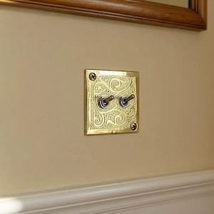 Op de afbeelding: Een messing lichtschakelaar met twee tuimelschakelaars. De vierkante plaat heeft een sierlijk, wervelend ontwerp. De schakelaar is gemonteerd op een beige muur, met een houten frame erboven.