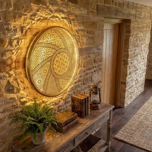 Puede incluir: Una decoración mural circular dorada con un intrincado diseño calado está montada en una pared de piedra texturizada. Debajo de la obra de arte hay una consola de madera con libros, una linterna y una planta en maceta.