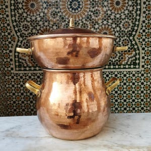 Pode incluir: Conjunto de panelas de cobre de duas peças com alças de latão. A panela superior tem uma tampa e fica em cima de uma panela maior. O cobre tem uma textura martelada, refletindo a luz. O fundo apresenta um design de mosaico.