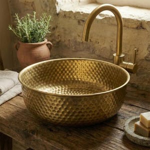 Op de afbeelding: Een gehamerde messing wastafel met bijpassende kraan. De wastafel is rond en staat op een houten oppervlak. Een kleine terracotta pot met een groene plant is zichtbaar op de achtergrond. Zeep en een klein schaaltje staan rechts.