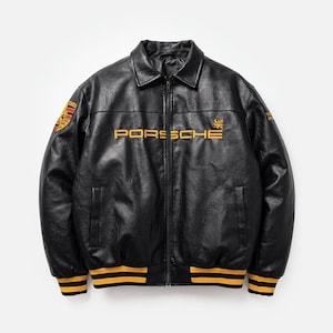 May include: A black leather jacket featuring a front zipper closure, a classic collar, and ribbed cuffs and hem with yellow stripes. The chest displays the yellow text PORSCHE, while the sleeves feature embroidered logos and emblems.