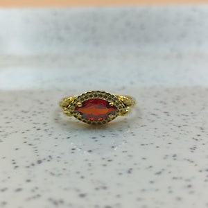 May include: A gold-colored ring with an oval, reddish-orange gemstone. The ring has a textured band and a detailed setting around the stone. The gemstone is the focal point, set in a detailed, ornate design.