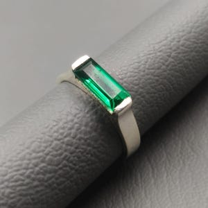 May include: A silver ring with a rectangular, emerald-green gemstone. The gemstone is set in a simple, modern band. The ring is displayed on a dark grey surface, highlighting the vibrant green of the stone.