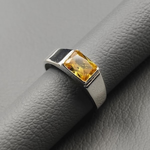 May include: A silver ring featuring a rectangular, yellow gemstone. The ring has a contemporary design with a polished finish. The gemstone is set in a bezel setting, and the band is smooth and simple.