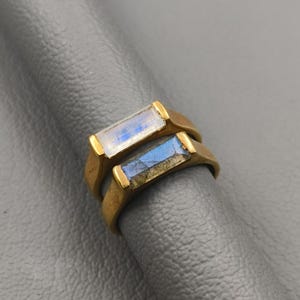 Puede incluir: Dos anillos dorados apilados uno encima del otro. Cada anillo presenta una piedra rectangular. Una piedra es azul pálido y blanca, la otra es azul y gris. Los anillos están sobre una superficie gris.