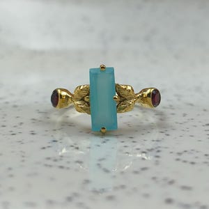 Może przedstawiać: Złoty pierścionek z prostokątnym, jasnoniebieskim kamieniem pośrodku i dwoma czerwonymi, okrągłymi kamieniami. Pierścionek ma wzór liści na obrączce. Biżuteria.